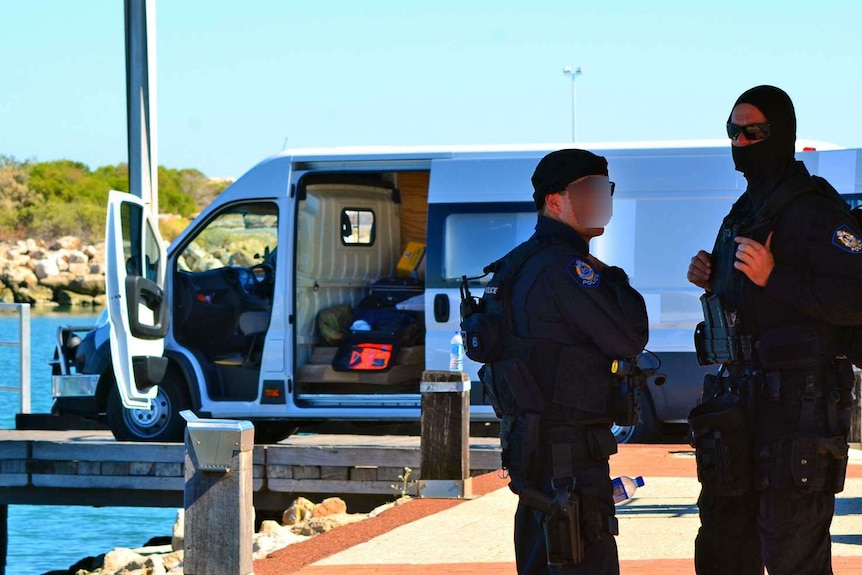 Two police officers, one wearing a balaclava, stand in front of a van with its door open parked on a jetty.