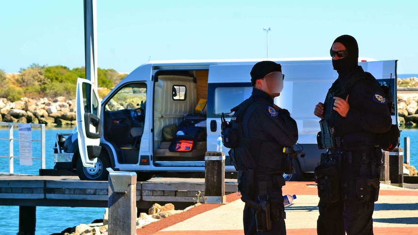 Two police officers, one wearing a balaclava, stand in front of a van with its door open parked on a jetty.