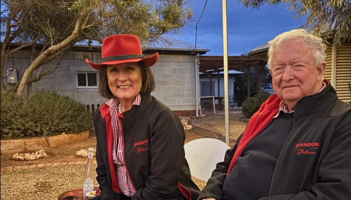 Una pareja sonríe a la cámara y viste chaquetas negras. La mujer lleva un sombrero rojo estilo vaquero.