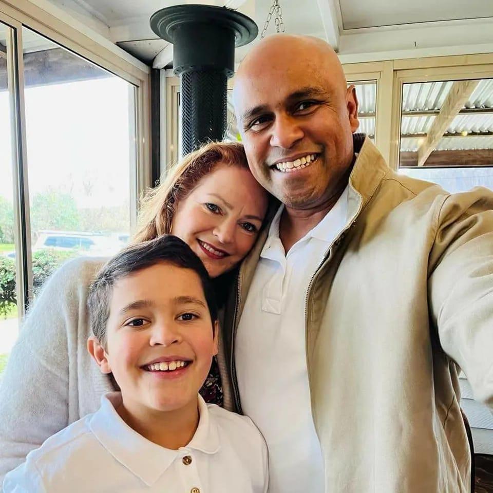 The family stands in a close huddle smiling with Emma resting her head on Raj's shouder 