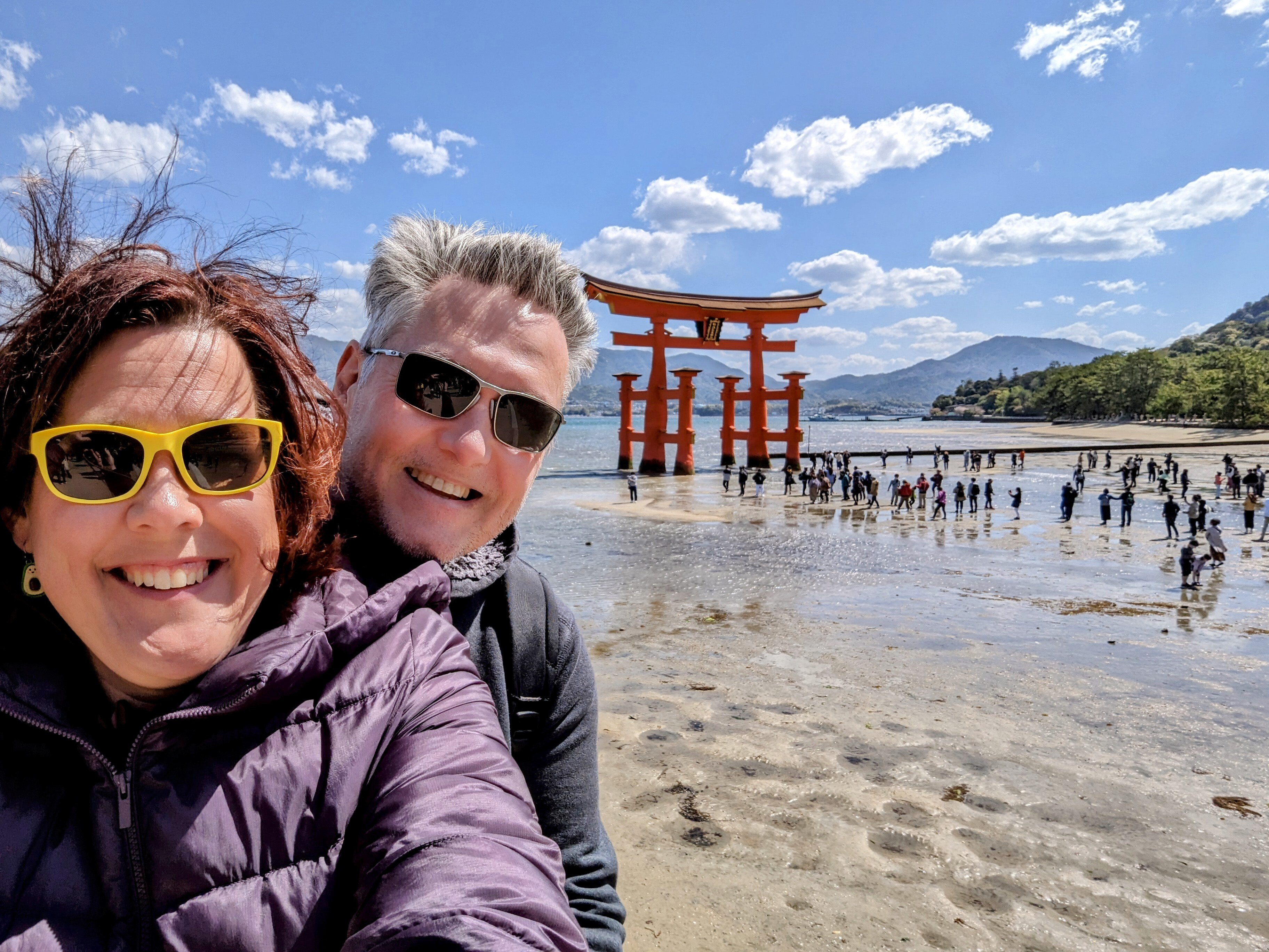 A woman with brown hair and yellow sunglasses takes a selfie with her partner at Japan's Itsukushima shrine.