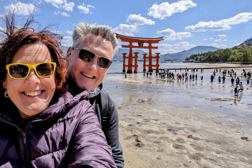 A woman with brown hair and yellow sunglasses takes a selfie with her partner at Japan's Itsukushima shrine.