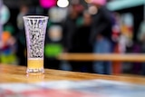 A half empty beer glass sits on a wooden table top beneath lights