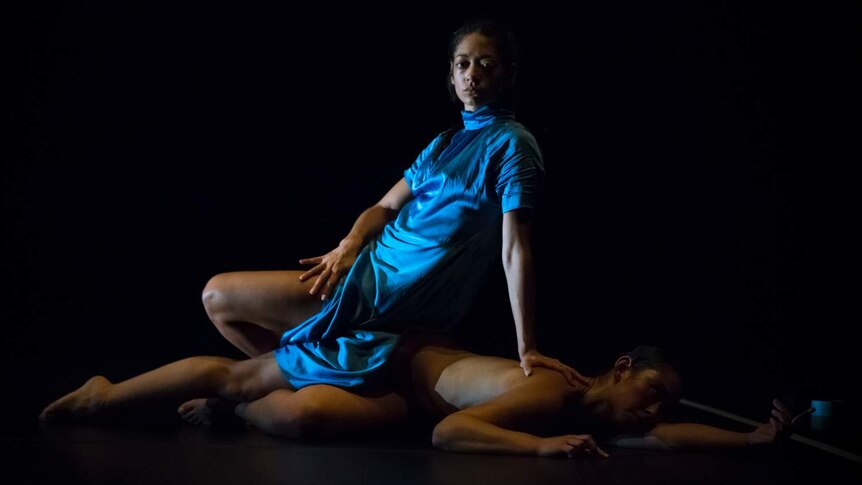 Woman in blue robe sits on body of other woman, naked, lying on stage floor.