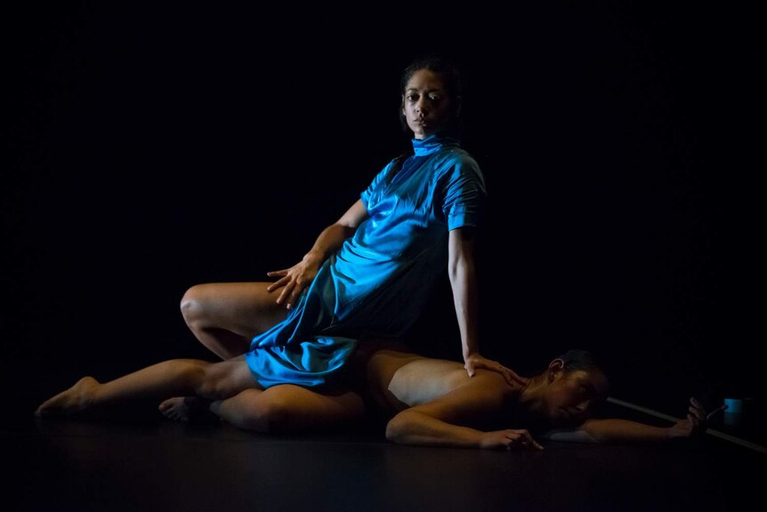 Woman in blue robe sits on body of other woman, naked, lying on stage floor.