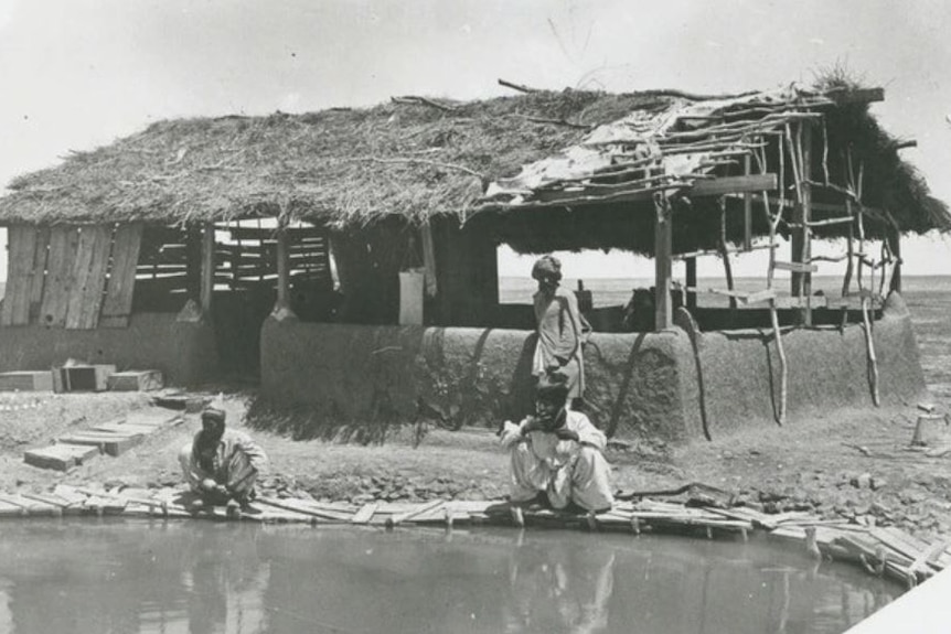 Black and white grainy photo of three Arabic-appearing men before a thatched roof hut near small body of water.