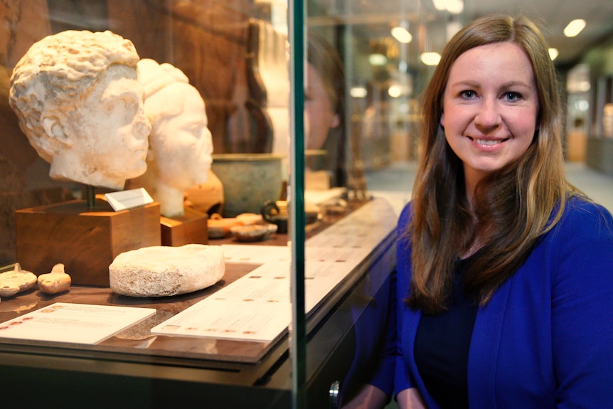 A woman with long brown hair stands in front of a display case containing a marble bust.
