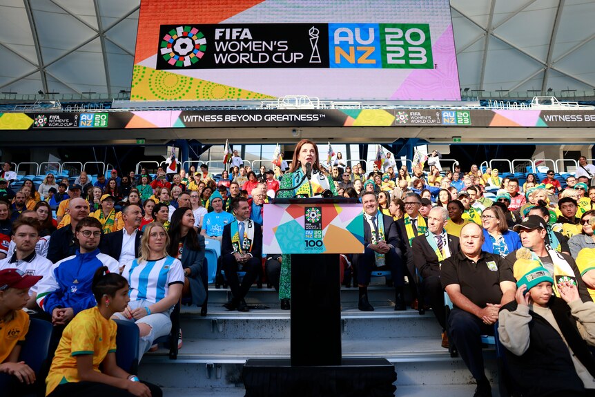 A woman in a green dress speaks at a podium surrounded by colourful fans and a big banner for a sports tournament