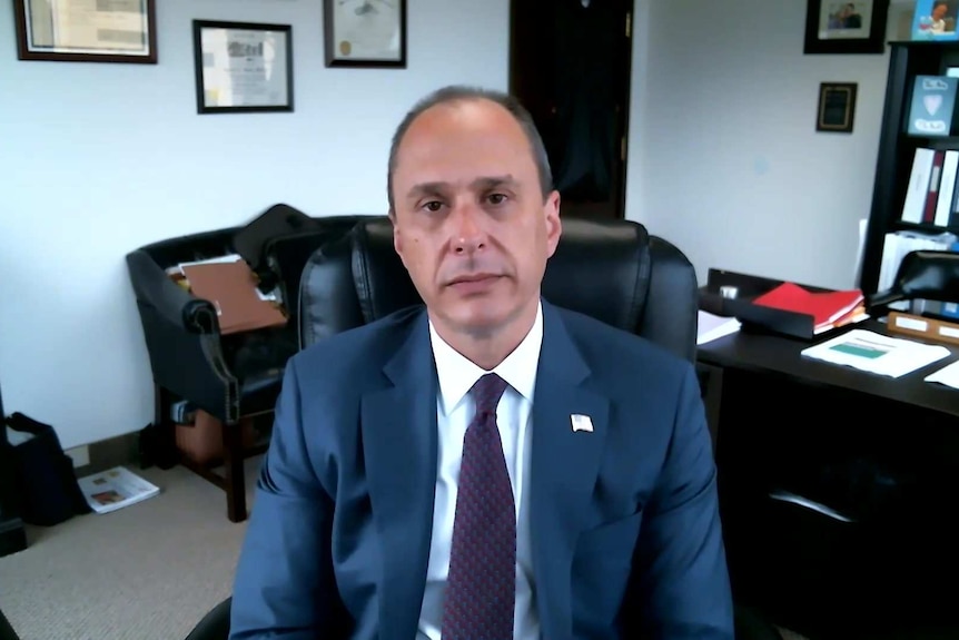 Spencer Kuvin sitting at his desk wearing a grey suit, white shirt and red-blue tie