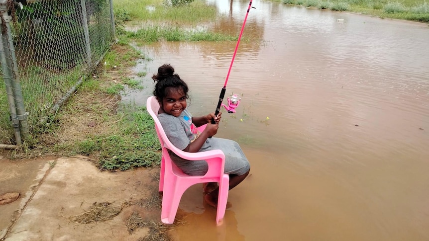 A small girl sits smiling in a chair fishing in floodwaters