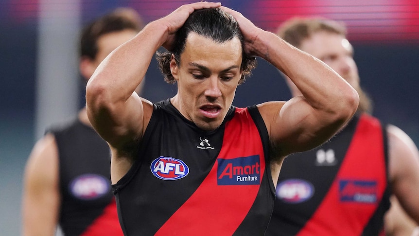 Dylan Shiel holds his hands behind his head and looks down towards the ground wearing a black singlet with a red sash