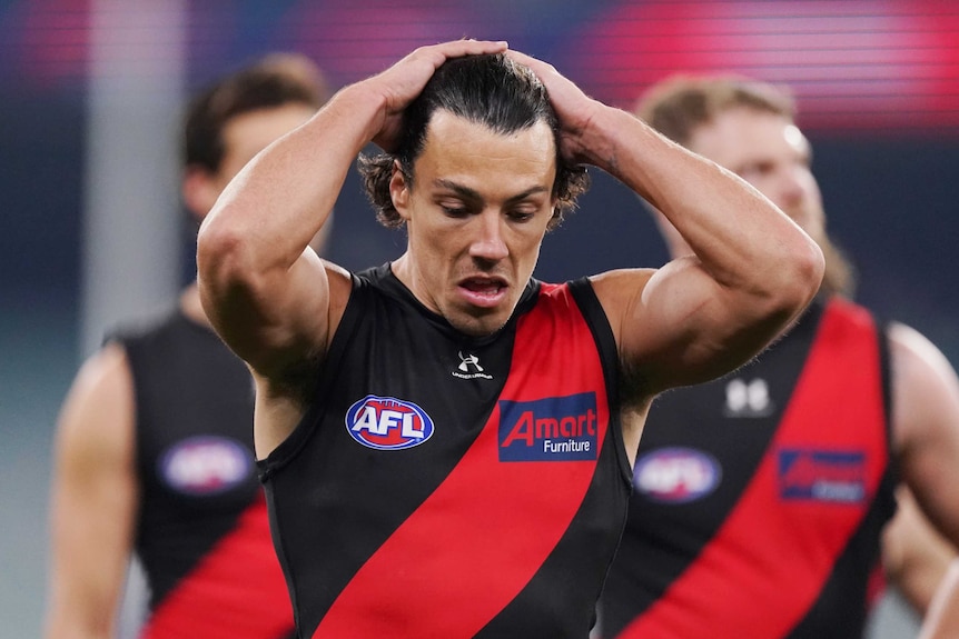 Dylan Shiel holds his hands behind his head and looks down towards the ground wearing a black singlet with a red sash