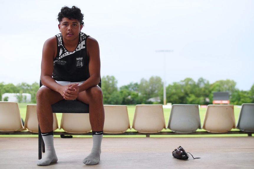 A boy looks at the camera, seated, with a single football boot on the ground next to him