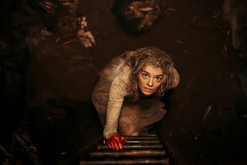 Samara Weaving looks up with fearful expression while climbing ladder with blood on her hand, with dirty hair and wedding dress.