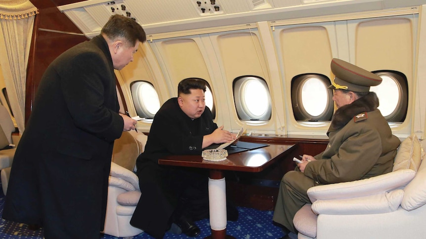 Kim Jong-un sits at a desk talking to an official onboard a plane.