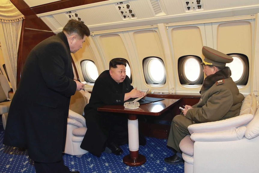 Kim Jong-un sits at a desk talking to an official onboard a plane.