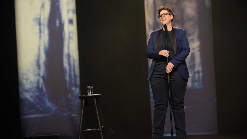 Gadsby in a navy and black suit stands onstage at microphone with abstract blue and white designs projected behind her.