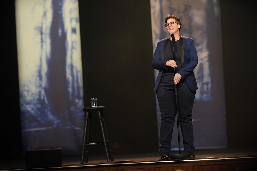 Gadsby in a navy and black suit stands onstage at microphone with abstract blue and white designs projected behind her.