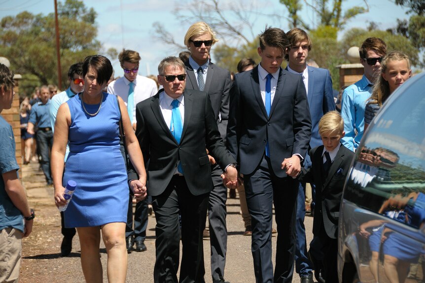 A mother and a father hold hands at the funeral of their son.