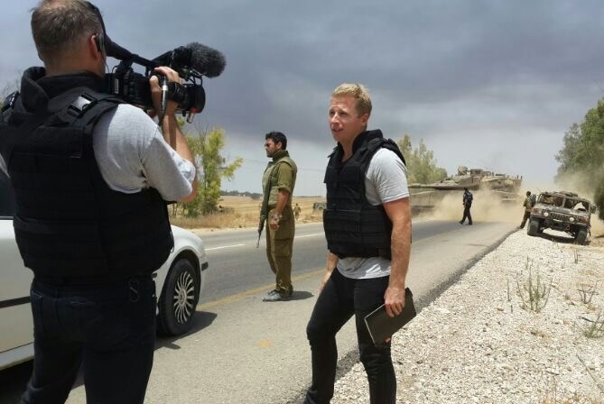 Macdonald wearing safety vest talking to camera with tanks and solders in background, location unknown.