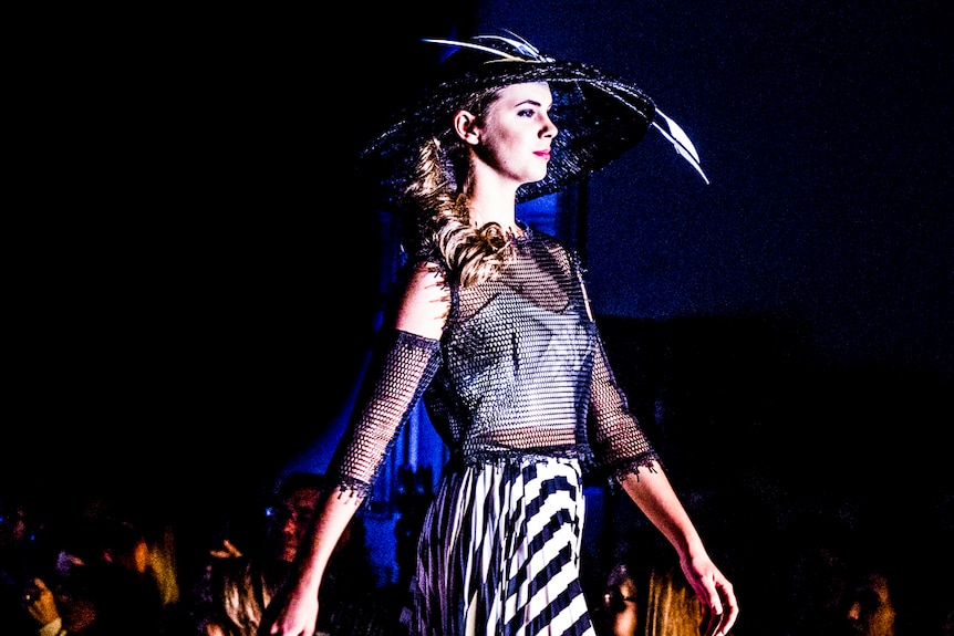 A teen model wearing a hat, black and white striped skirt, and mesh top walks down the runway, in front of a dark background.