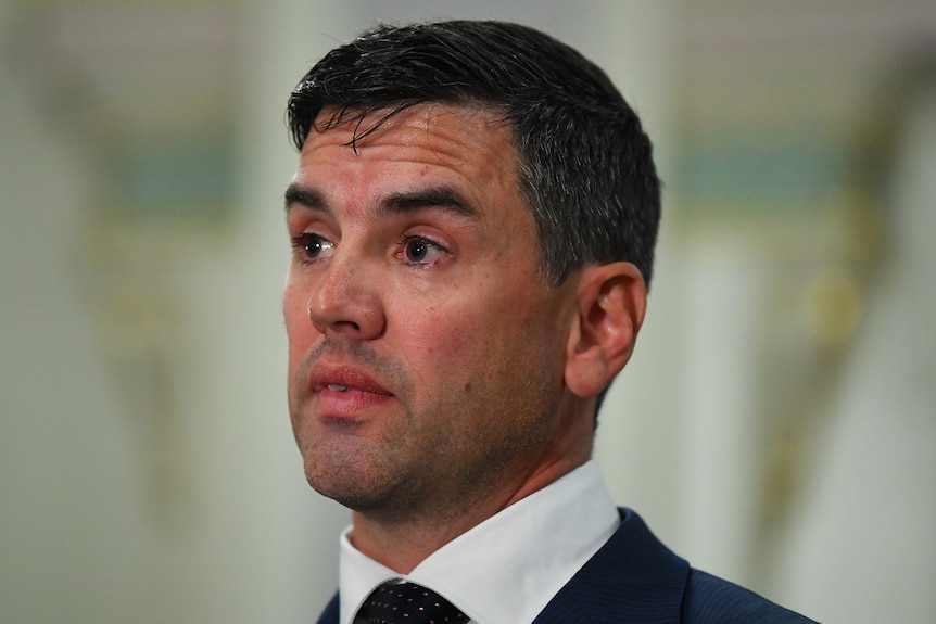 Brad Battin stands indoors, dressed in a white shirt and jacket as he holds a press conference.