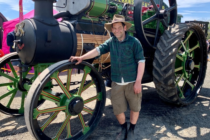 A man in a hat and green plaid shirt stands in front of a shining green and black steam engine.