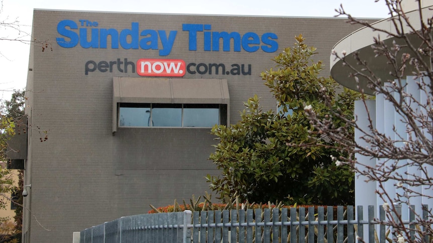 A greay brick building stands behind a tree with logos for The Sunday Times and perthnow.com.au on its wall.