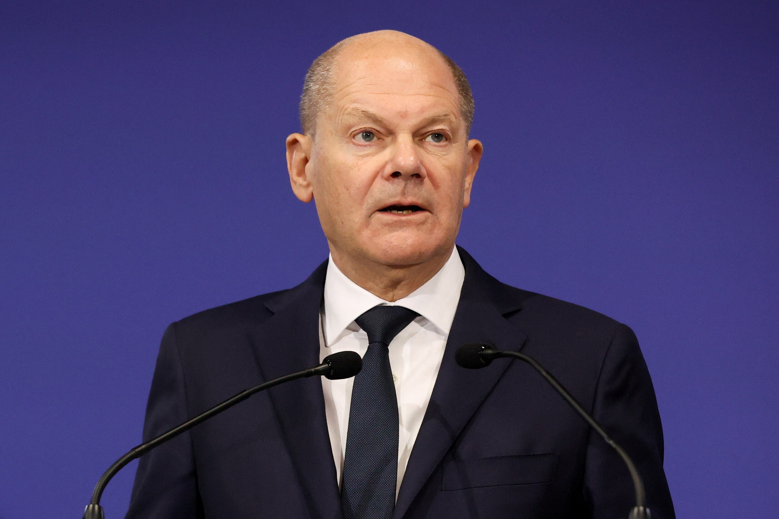 Olaf Scholz wearing a navy suit standing in front of two microphones.