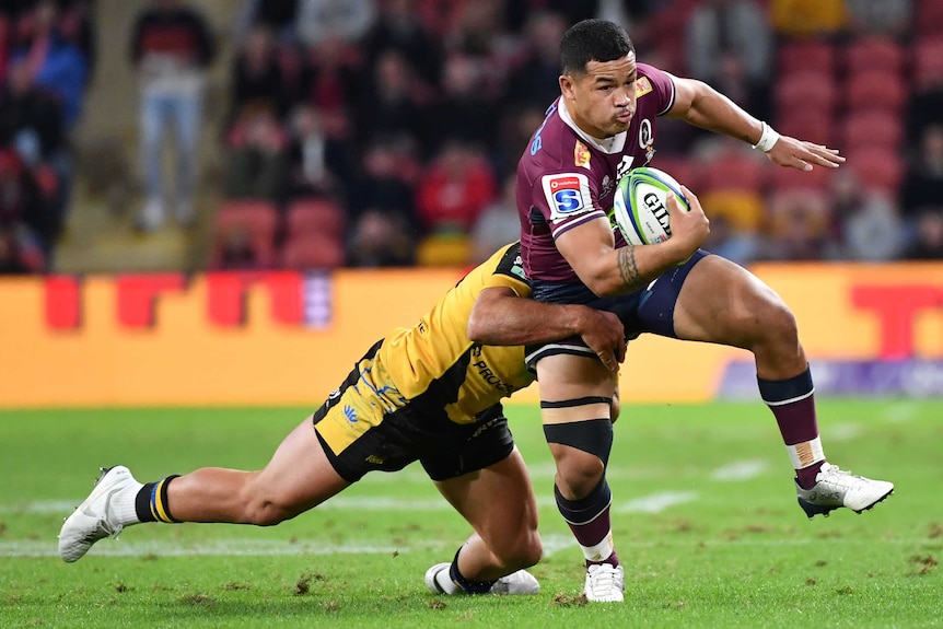 A Queensland Super AU player holds the ball with his right arm as he is tackled around his thighs by a Western Force opponent.