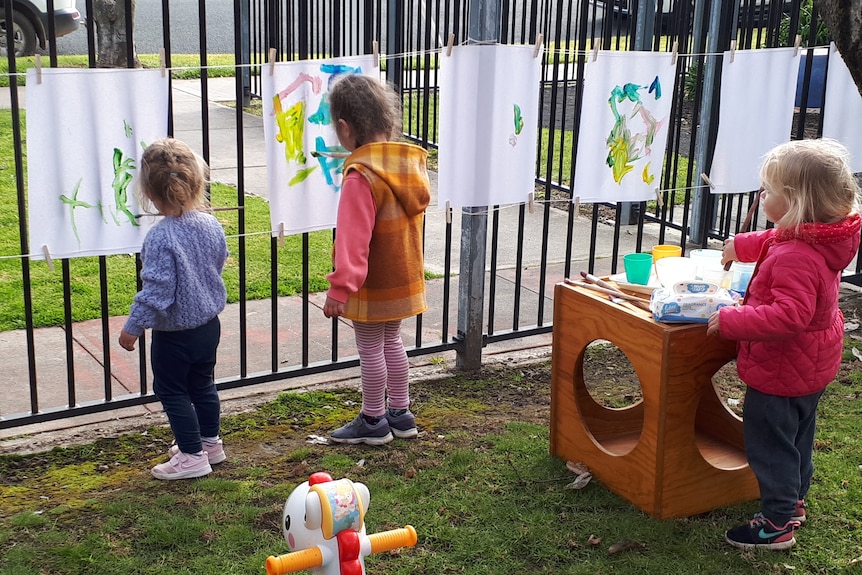 Three young girls stand with their backs to the camera and paint on paper attached to a fence.