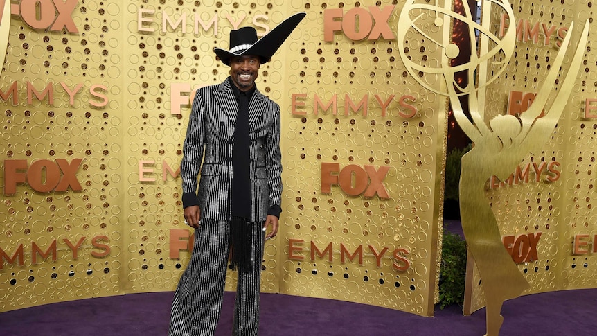 Billy Porter wears a statement black hat and black and white pinstripe suit as he stands against a gold background.