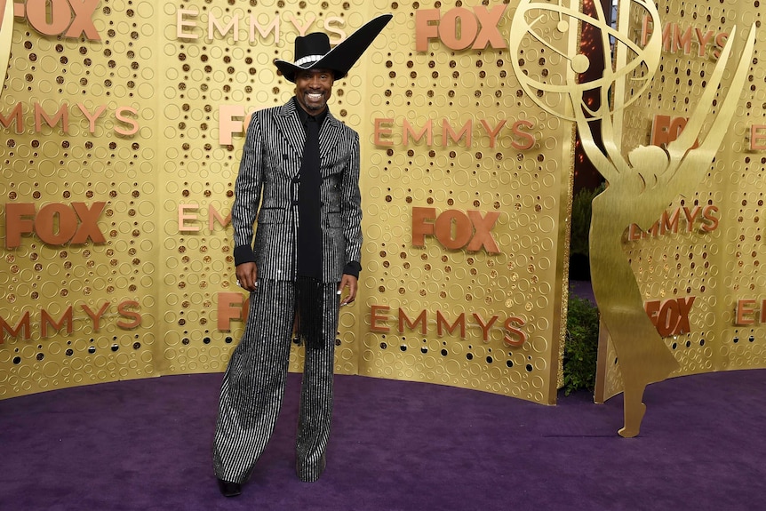 Billy Porter wears a statement black hat and black and white pinstripe suit as he stands against a gold background.