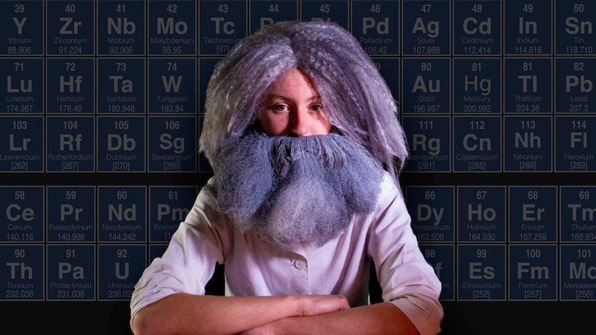 A young boy dressed up with fake beard and hair to look like a scientist, the periodic table in the background.