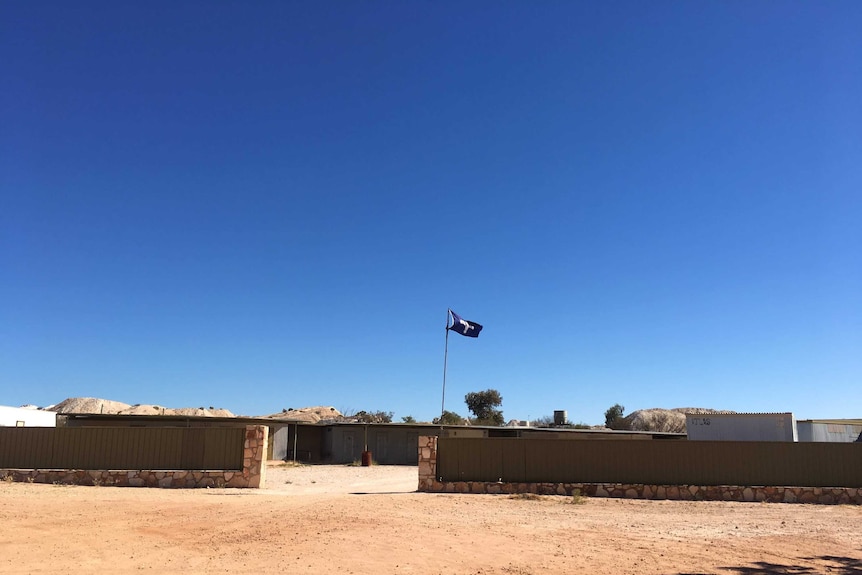 A Eureka stockade flag flies in front of a transportable grey home on a green fence with piles of rubble in background