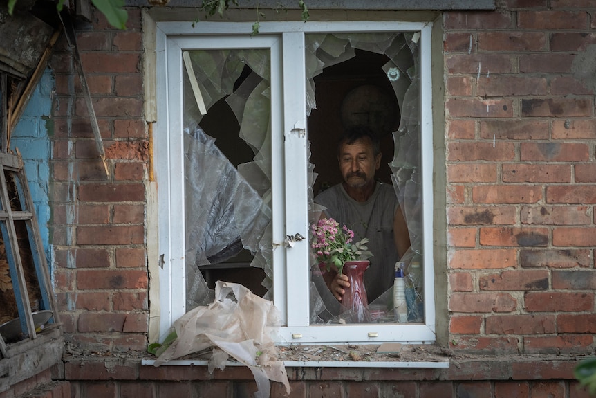 A man with a mustache is pictured through a broken window as he places a vase with flowers in it. 
