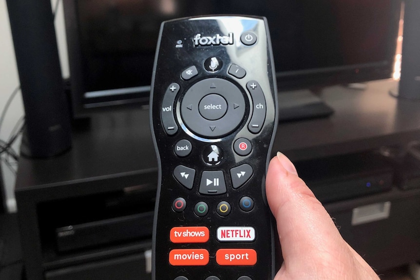 A hand holding a Foxtel remote in front of a television.
