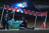 Police and doctors under a marquee behind a sheet on the beach at night.