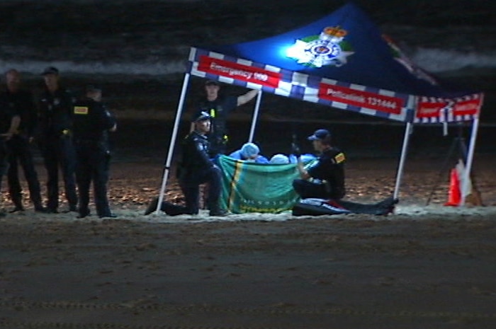 Police and doctors under a marquee behind a sheet on the beach at night.