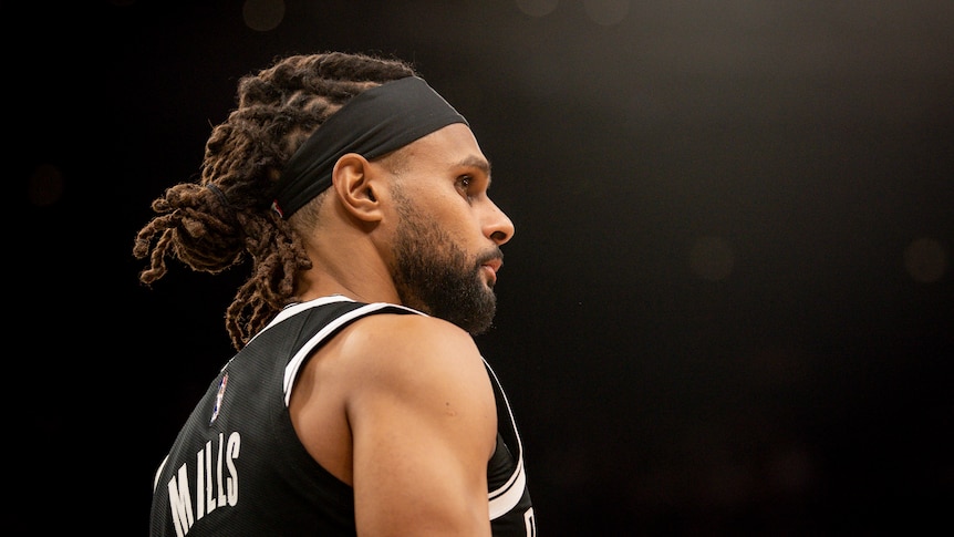 A side-on, low view of NBA player Patty Mills with a black background behind him before a Brooklyn Nets game.