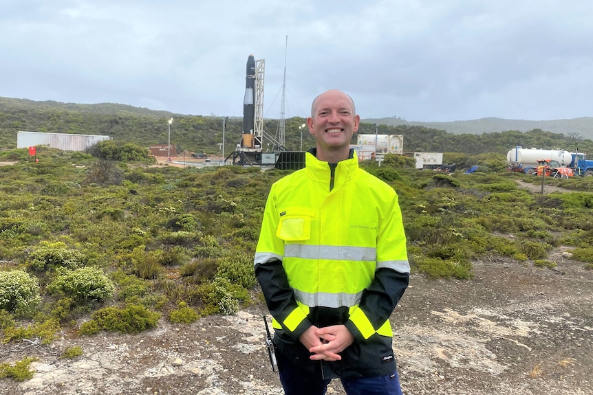 Smiling bald man in hi-vis jacket standing in front of rocket in background