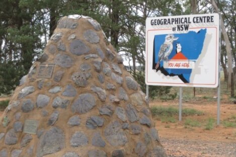 A large pile of slag and stones with a plaque on it in front of a sign saying 'geographical centre of NSW'.