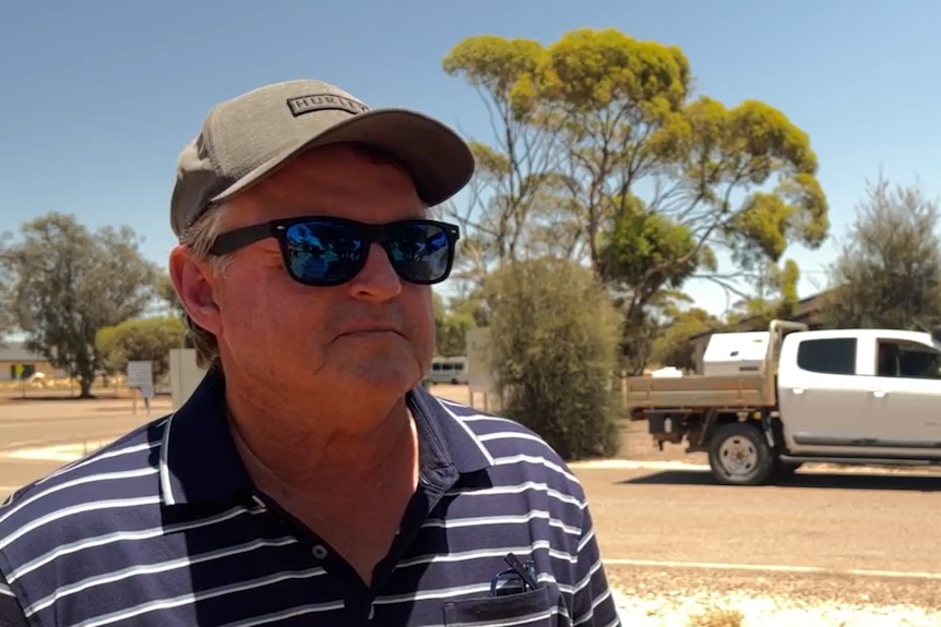 A man in a hat and dark sunglasses with a ute and gum tree behind him