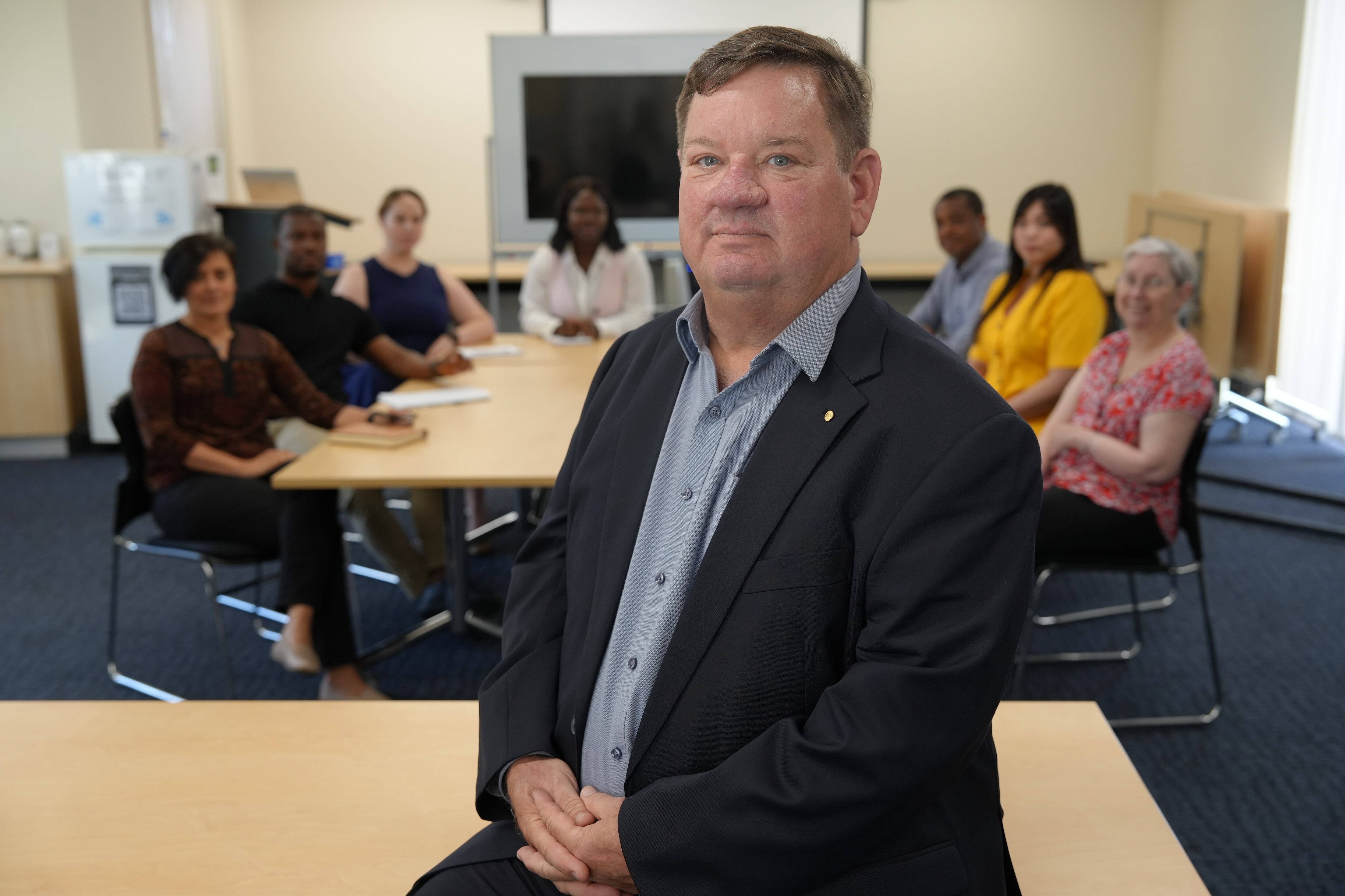 A man in a suit sits in front of a round table of people in professional attire.