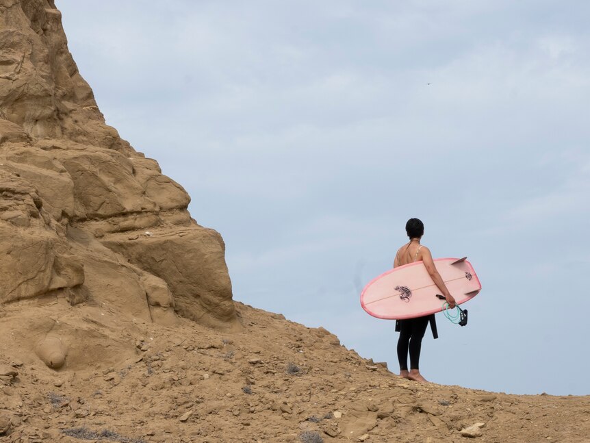 A woman surfer stands on a rocky mountain, holding her surfboard, looking in the distance. 