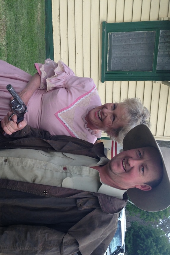 Woman in pink frilly colonial style dress, laughing with man dressed in heavy outdoor clothes and hat, holding a pistol. 