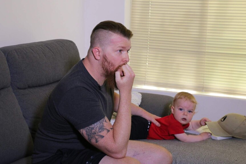 Nicholas and his toddler son James sitting on a couch, with Nicholas holding his hand to his mouth with a concerned expression.