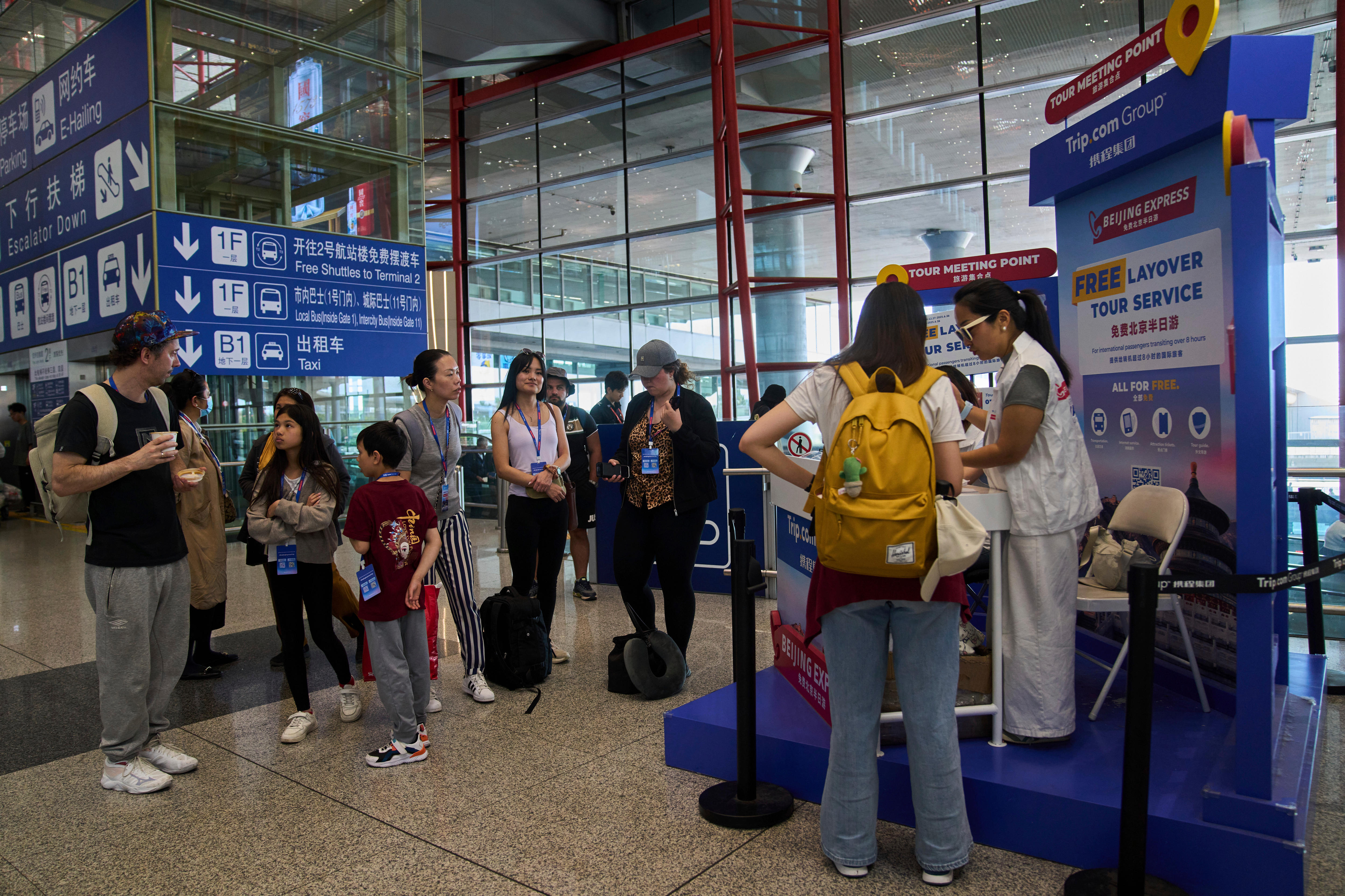 tourists including two children carrying backpacks wait at the airport while a woman with yellow backpack talks to tour guide