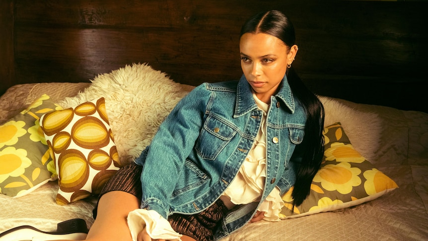 TSHA sits on a bed with yellow patterned cushions, wearing a denim jacket, brown skirt & slick ponytail.