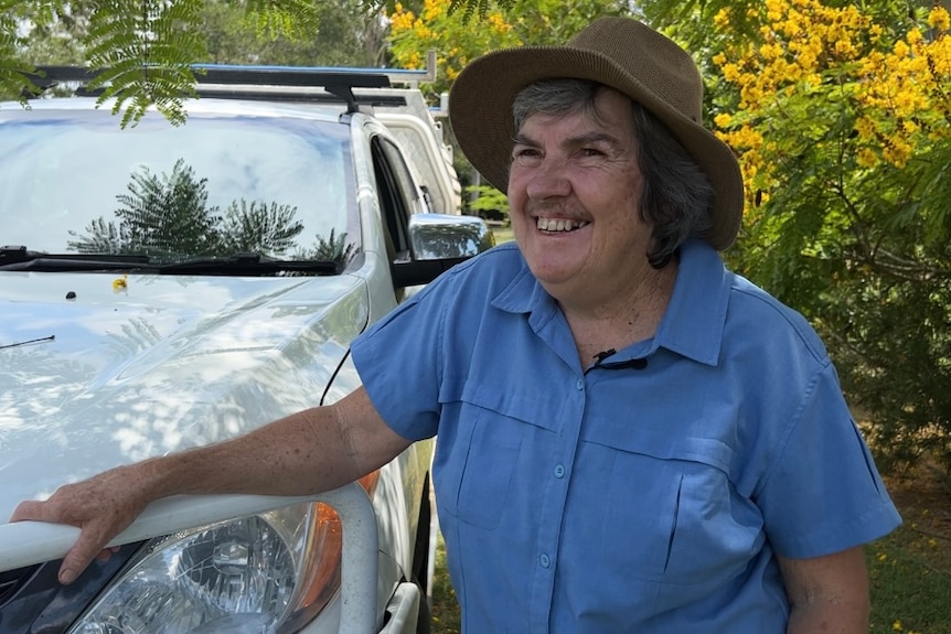 A woman in a blue shirt and hat smiles while leaning against a white care